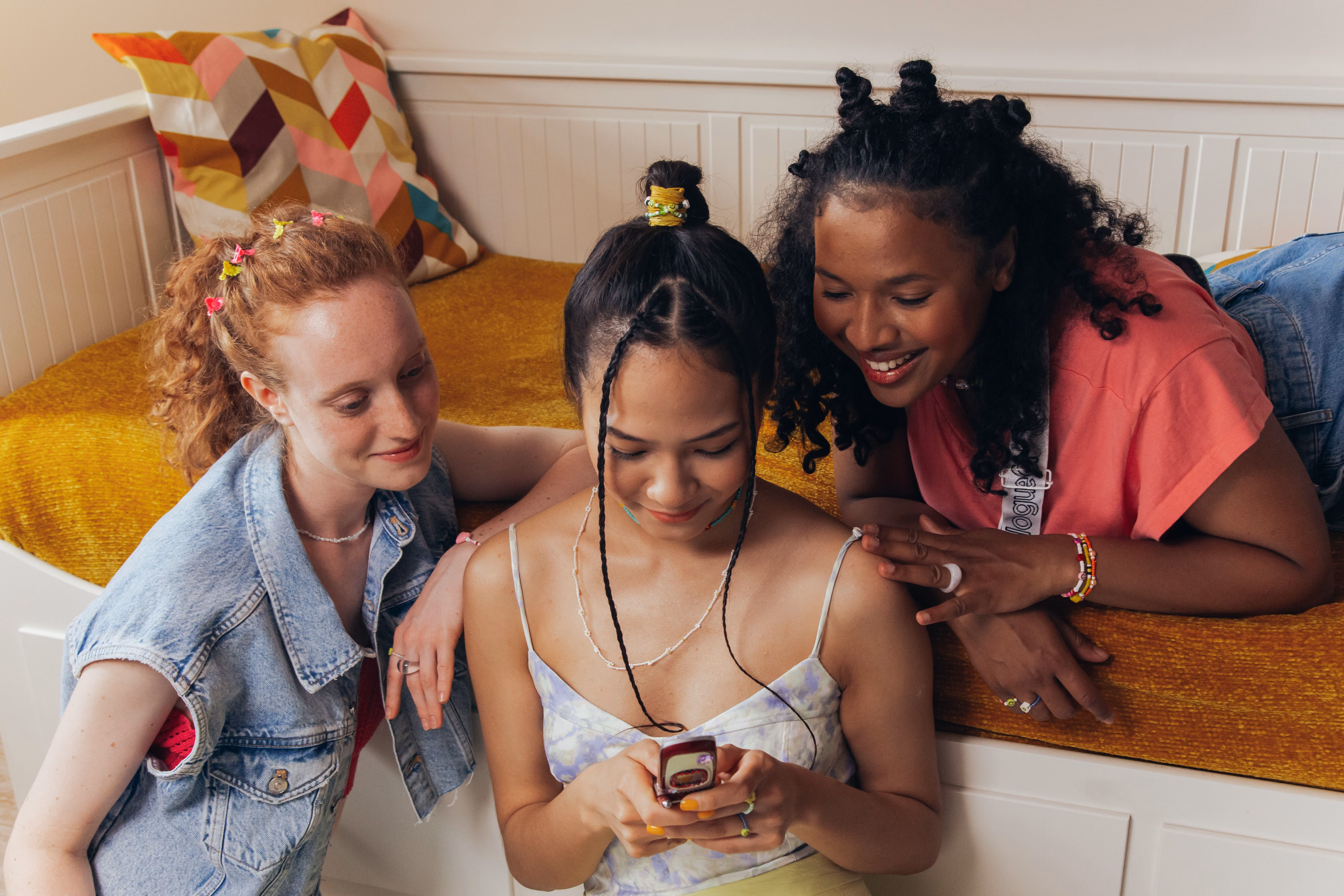 Trois jeunes femmes souriantes regardant un téléphone portable assises près d'un lit avec une couverture jaune.